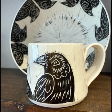 Handmade by pots by DJR this porcelain mug was hand thrown and hand carved using sgraffito techniques. Porcelain is white with black glaze carved away to create images of two ravens sitting together. This image shows one side of the coffee mug sitting in a made in Buffalo gift shop in Buffalo, NY