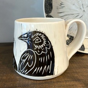 Handmade by pots by DJR this porcelain mug was hand thrown and hand carved using sgraffito techniques. Porcelain is white with black glaze carved away to create images of two ravens sitting together. This image shows one side of the coffee mug sitting in a made in Buffalo gift shop in Buffalo, NY