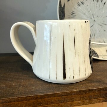 Handmade by pots by DJR this porcelain mug was hand thrown and hand carved using sgraffito techniques. Porcelain is white with black glaze carved away to create images of two ravens sitting together. This image shows the back side of the coffee mug sitting in a made in Buffalo gift shop in Buffalo, NY