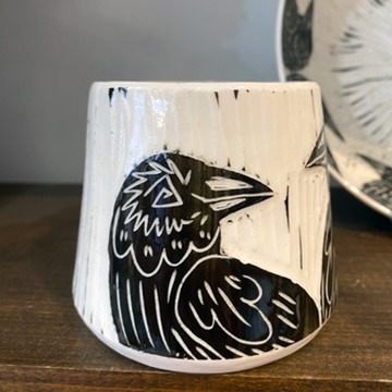 Handmade by pots by DJR this porcelain mug was hand thrown and hand carved using sgraffito techniques. Porcelain is white with black glaze carved away to create images of two ravens sitting together. This image shows one side of the coffee mug sitting in a made in Buffalo gift shop in Buffalo, NY